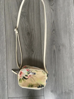 Blommig axelväska från Lycke - Snygg axelväska från Lycke i beige med blommigt mönster i grönt, rosa och blått, samt en fin ljusgul bakgrund. Väskan har justerbar axelrem och en rektangulär form. Perfekt för dig som vill ha en liten och smidig väska med tropisk vibe.