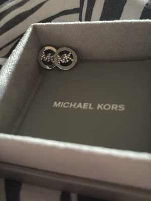 Silverfärgade örhängen Michael Kors - Säljer ett par snygga silverfärgade örhängen från Michael Kors. De är runda med glittrande stenar i mitten och har MK-loggan som detalj. Materialet är metall och designen är stilren med en lyxig touch.