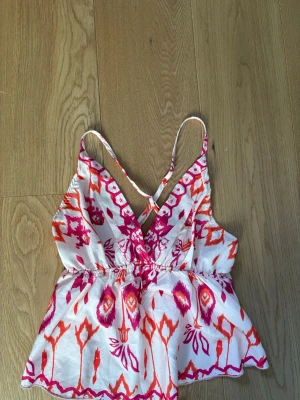 Vit och rosa mönstrad linnetopp - Säljer en snygg linnetopp med färgglatt mönster i rosa och orange på vit botten. Toppen har smala axelband som korsas i ryggen och en v-ringad front. Perfekt för varma dagar och sommarens outfits.