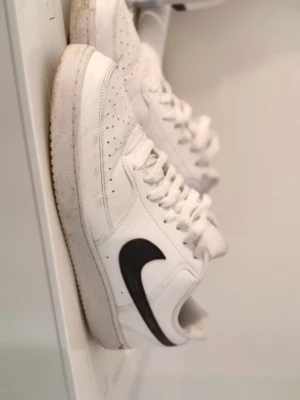 Vita Nike sneakers med svart Swoosh - Klassiska vita Nike sneakers med svart Swoosh på sidan. Skorna har lågt skaft, perforerade detaljer vid tårna och vit sula. Tillverkade i läder med textilfoder och snörning framtill. Perfekta för en clean och sportig look. Använd 5 ggr