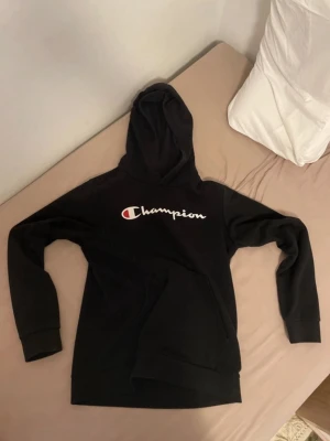 Champion Hoodie - Svart hoodie från Champion med stor vit och röd logga på bröstet. Klassisk modell med huva och känguruficka framtill. Perfekt för en avslappnad och sportig stil. Mjuk bomullsblandning som är skön att ha på sig.
