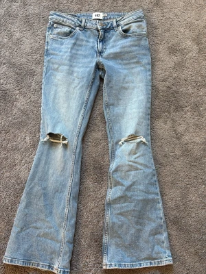 Ljusblå bootcut jeans med slitningar - Säljer ett par ljusblå bootcut jeans med slitna detaljer på knäna. Jeansen har klassisk femficksdesign, normal midja och är tillverkade i denim. Perfekta för dig som gillar en avslappnad och trendig look.