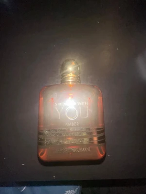 Armani Stronger With You Amber parfym - Upptäck Stronger With You Amber från Giorgio Armani, en exklusiv parfym i en elegant, fyrkantig glasflaska med gyllene lock och bärnstensfärgad vätska. Flaskan har stilrena guldiga detaljer och text. Perfekt för dig som gillar lyxiga dofter.