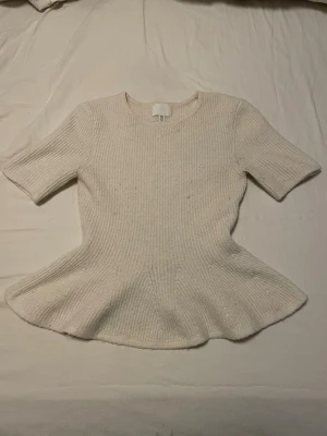 Vit ribbad topp med volang nedtill  - Säljer en kräm vit ribbad topp från H&M med volang liknande nedtill. Toppen har rund halsringning och korta ärmar. Skriv om fundering hur den ser ut på! Knappt använd 