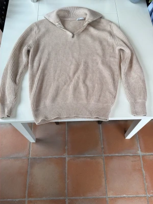 Beige ribbstickad tröja med dragkedja - Säljer en beige ribbstickad tröja med krage och kort dragkedja framtill. Tröjan har långa ärmar och är tillverkad i ett mjukt, tjockare material som passar perfekt för kyligare dagar. Enkel och stilren design som är lätt att matcha.