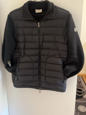 Svart cardiganjacka från Moncler - Snygg svart cardiganjacka från Moncler med quiltad framsida och mjuka stickade ärmar. Jackan har dragkedja framtill, två sidofickor med dragkedja och en liten Moncler-logga på ärmen. Perfekt för lager-på-lager och streetwear.