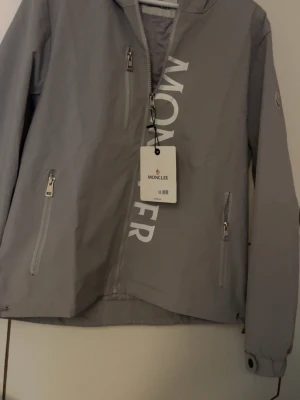 Grå vindjacka  - Snygg grå vindjacka  med vit logotyp på framsidan. Jackan har huva, tre dragkedjeförsedda fickor och justerbara ärmslut. Tillverkad i lätt och slitstarkt material