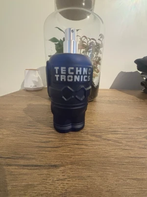 Technotronics blå parfymflaska - Cool parfymflaska från Technotronics i matt mörkblå plast med futuristisk design och silverfärgad spraytopp. Flaskan har ett unikt, nästan robotliknande utseende och tydlig logga framtill. Perfekt för dig som gillar modern och edgy stil.