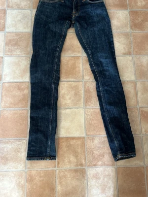 Mörkblå jeans från Nudie Jeans - Säljer ett par mörkblå jeans från Nudie Jeans med klassisk femficksmodell och snygga kontrastsömmar. Bakfickorna har Nudie Jeans ikoniska broderade vågmönster i orange. Jeansen har smal passform och är tillverkade i denim.
