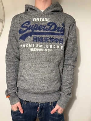 Grå Superdry hoodie med blått tryck - Säljer en grå hoodie från Superdry med blått och vitt tryck på bröstet där det står 'Vintage Superdry Premium Goods' samt japanska tecken. Hoodien har huva, magficka och ribbade muddar. Perfekt för en avslappnad streetwear-look. Pris kan diskuteras!💸 Strl: M passar S✅ Hör av dig om du har funderingar!👍