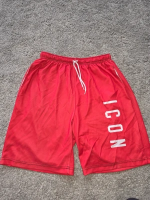 Röda ICON shorts med snörning - Snygga röda shorts från ICON med vit snörning i midjan och tryckt text längs benet. De har en avslappnad passform och är perfekta för sommardagar. Materialet känns som polyester och shortsen har elastisk midja för extra komfort.