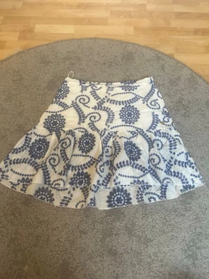 Vit och blå blommig kjol från By Malina - Säljer en fin kjol från By Malina med vit bas och blått blommigt mönster. Kjolen har volang nertill och en liten guldfärgad detalj på sidan. Materialet är lätt och luftigt, perfekt för varma dagar. Passformen är normal och kjolen har en  trendig look💓aldrig använd💓