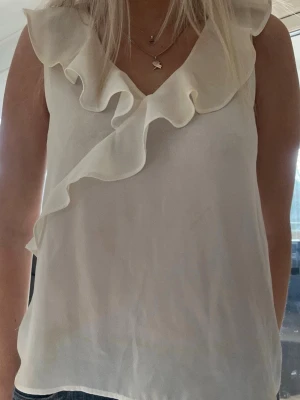 Vit ärmlös blus med volang - Säljer en stilren vit blus med volangdetaljer längs ringningen och framtill. Blusen är ärmlös och har en lätt, luftig känsla i ett tunt tyg som ger ett elegant intryck. Perfekt att matcha med jeans eller kjol för en trendig look.