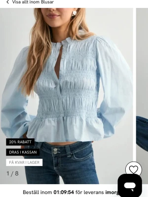 Ljusblå smockad blus med volang - Trendig ljusblå blus från Nelly med smockade detaljer över bröst och midja, volang nedtill och långärmade puffärmar. Ståkrage och knappöppning framtill ger en chic look. Perfekt att matcha med jeans för en cool stil.