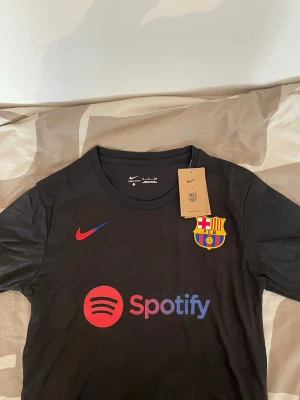 Svart FC Barcelona matchtröja Nike - Säljer en svart FC Barcelona matchtröja från Nike med klubbens emblem på bröstet och Spotify-logga i rött och lila. Tröjan har rund hals och diskreta mönster av klubbens logga i tyget. Perfekt för dig som vill representera Barca på planen.