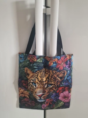 Tygväska leopardmönster unisex - Tygväska för unisex. Väskan är tillverkad i polyester och har ett färgstarkt tryck med leopardmotiv och blommor på framsidan och baksidan.Utrustad med två axelremmar i tyg för bärning över axeln eller i handen. Passar för vardagsbruk, exempelvis som shoppingväska eller extra vardagsväska. 38 cm× 33,5 cm.