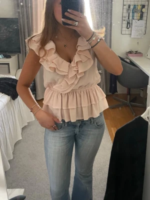 Ljusrosa volangblus med v-ringning - Supersöt ljusrosa blus med v-ringning och stora volanger framtill. Blusen är ärmlös och har en elastisk midja som ger en snygg passform. Perfekt för dig som gillar romantiska detaljer och en feminin look.