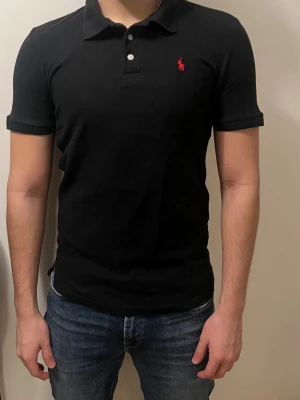 Svart pikétröja från Polo Ralph Lauren - Klassisk svart pikétröja från Polo Ralph Lauren med röd broderad logga på bröstet. Tröjan har korta ärmar, krage och knäppning med två vita knappar. Perfekt till jeans eller shorts för en clean och stilren look. Storlek m/s