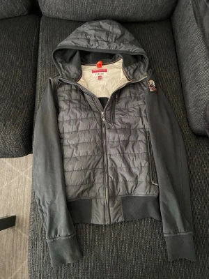 Svart cardigan från Parajumpers - Snygg svart cardigan från Parajumpers med quiltad front, huva och dragkedja. Jackan har ett broderat märke på ärmen och mjuka muddar vid ärmslut och nederkant. Insidan är fodrad med beige fleece för extra komfort. Perfekt för blåsiga dagar.