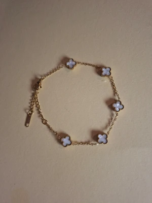 Guldfärgat armband med vita blommor - Snyggt armband i guldfärgad metall med fem vita fyrklöverformade detaljer. Kedjan är tunn och smidig med justerbar längd. Perfekt accessoar för att lyfta din outfit med en elegant touch.
