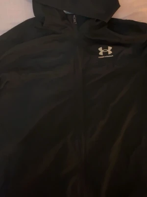 Svart vindjacka från Under Armour - Snygg svart vindjacka från Under Armour med huva och dragkedja framtill. Jackan har en sportig look och är tillverkad i lätt syntetmaterial med meshfoder för extra komfort. Perfekt för aktiva dagar och träning utomhus.