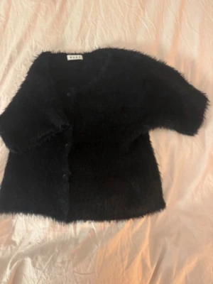 Svart fluffig kofta med knappar - Mysig svart kofta med fluffig textur och knäppning framtill. Koftan har korta ärmar och rund halsringning, perfekt för dig som gillar en trendig och unik look. Passar till många olika stilar och är superenkel att matcha.