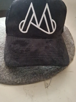 Svart truckerkeps med vit logga - Snygg svart truckerkeps med vit broderad logga framtill. Kepsen har böjd skärm och mesh på sidorna och bak för extra ventilation. Justerbar passform med snapback-spänne. Perfekt för en avslappnad streetwear-look.