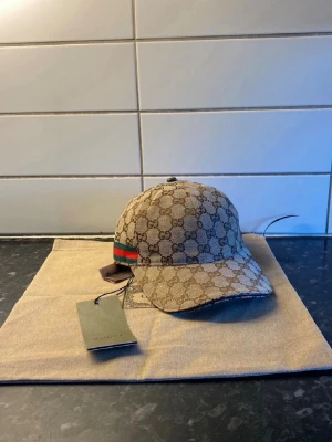  beige keps med GG-mönster - Säljer en beige keps med det klassiska GG-monogrammönstret över hela kepsen. På sidan finns en grön och röd rand som signaturdetalj. Kepsen är i canvasmaterial och har justerbart band bak. Kommer med originalpåse och taggar.