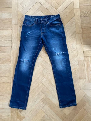Archieve jeans med slitningar - Säljer ett par mörkblå archieve stil jeans med slitningar vid knäna. Jeansen är från ett okänt märke och är i väldigt fint skick. Storlek W32/L32. Är öppen för prisförslag och frågor om jeansen.