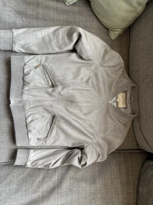Silver bomberjacka från River Island - Snygg silverfärgad bomberjacka från River Island med ribbade muddar och krage. Jackan har två snedställda fickor med dragkedja och stängs med dragkedja framtill. Materialet har en lätt glansig finish som ger en cool look.