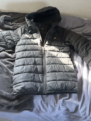 Svart pufferjacka från Lyle & Scott - Snygg svart pufferjacka från Lyle & Scott Junior med huva och dragkedja framtill. Jackan har det klassiska guldiga logomärket på bröstet och quiltad design. Perfekt för kalla dagar och riktigt skön att bära.
