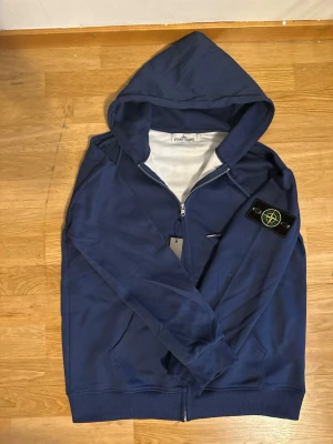Blå hoodie från Stone Island - Snygg blå hoodie från Stone Island med dragkedja framtill och klassisk logotyp-patch på ärmen. Tillverkad i mjukt bomullsmaterial och har två fickor fram. Perfekt för en avslappnad streetwear-look.