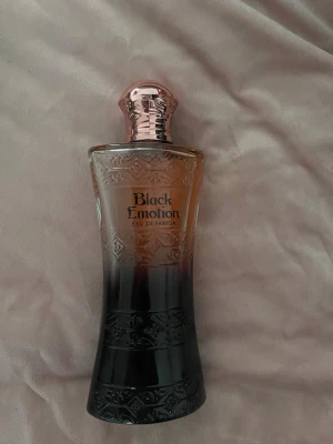 Black Emotion Eau de Parfum - Snygg parfymflaska från Black Emotion med elegant design i glas. Flaskan har en snygg färgskiftning från roséguld upptill till svart nedtill och ett mönstrat lock i metall-look. Perfekt för dig som gillar lyxiga och unika dofter.