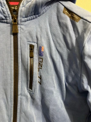 Blå Sail Racing kofta - Blå hoodie med dragkedja från Sail Racing. Har huva med snörning, bröstficka med dragkedja och tryck med australiensisk flagga och texten NZL 77. Mjukt material och patch på ena ärmen ger en sportig look. Koftan är i barnstorlek och har storleken 170 vilket motsvarar storlek S 
