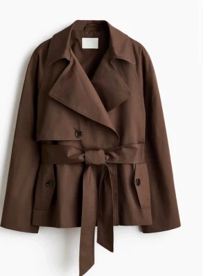 Brun trenchcoat från H&M med knytskärp - Snygg brun trenchcoat från H&M med breda slag, dubbelknäppning och knytskärp i midjan. Jackan har två fickor med knappstängning och är tillverkad i ett kraftigt material som ger en cool och modern look.