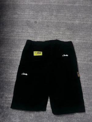 Svarta shorts från cortiez - Svarta shorts från Guerilla Group med vita logotryck och en gul patch på ena benet. Baksidan har en stor oval grafisk print i grått. Shortsen har en loose passform och är perfekta för en streetwear-look.