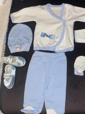 Sött babyset i blått och vitt - Ett gulligt babyset i ljusblått och vitt som innehåller byxor med fot, omlottkofta med blå rosett, mössa, vantar och små skor. Setet har mjukt material och fina detaljer som blå kantband och en liten applikation på mössan.
