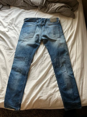 Blå Replay jeans med slitningar - Säljer ett par blå jeans från Replay med snygga slitningar och tvättade detaljer. Klassisk femficksmodell med raka ben och läderpatch bak i midjan. Jeansen har kontrastsömmar och en liten röd detalj på bakfickan. Perfekta för en avslappnad och trendig look. Och dem har ett hål i knät för extra snygg vintage look