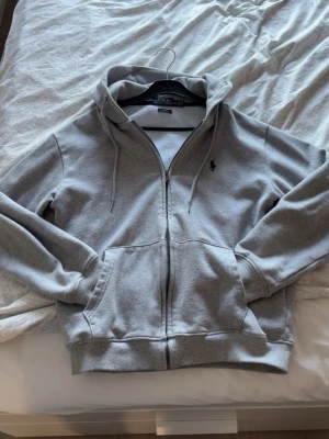 Grå zip-hoodie från Polo Ralph Lauren - Snygg grå hoodie med dragkedja från Polo Ralph Lauren. Klassisk design med huva, dragsnören och två fickor framtill. Liten broderad logga på bröstet. Perfekt för en chill och stilren look.