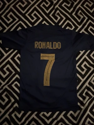 Blå Al Nassr Ronaldo matchtröja - Snygg mörkblå Al Nassr matchtröja med guldiga detaljer, Ronaldo och nummer 7 på ryggen. Tröjan har klubbmärke, Nike-logga och KAFD sponsortryck på bröstet. Tillverkad i lätt och ventilerande material, perfekt för fotboll eller träning.