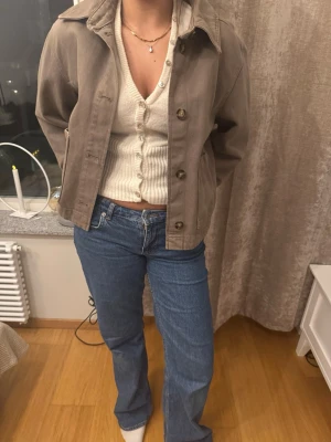 Beige jeansjacka med klassisk krage - Snygg beige jeansjacka med klassisk krage och knäppning framtill. Jackan har raka ärmar och en avslappnad passform som ger en cool och tidlös look. Perfekt att slänga över en stickad tröja eller t-shirt för en casual vibe.