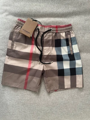 Rutig Burberry shorts med snörning - Säljer ett par klassiska Burberry shorts med det ikoniska rutiga mönstret i beige, svart, vit och röd. Shortsen har elastisk midja med svart snörning och fickor på sidorna samt en bakficka. Perfekta för sommaren och riktigt snygga till streetwear.