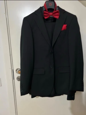 Svart Bal kostym regular fit - Säljer en stilren svart kostym med matchande svart skjorta, röd fluga och röd näsduk. Den har använts 1 gång under min bal så den är i perfekt skick. Den passar 165-170 ungefär. Pris kan alltid diskuteras! Allting ingår!