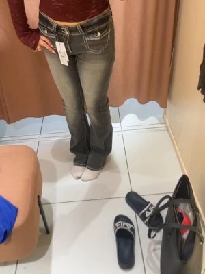 Grå bootcut jeans med kontrastsömmar - Säljer ett par grå bootcut jeans med vita kontrastsömmar och dekorativa fickor bak. Jeansen har låg midja och detaljerade sömmar både fram och bak. Perfekta för dig som gillar en retro och trendig look. 