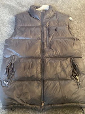 Polo Ralph lauren väst - Snygg grå dunväst från Polo Ralph Lauren med hög krage, dragkedja framtill och tre fickor med dragkedja. Klassisk broderad logga på bröstet och justerbara spännen i sidorna. Perfekt för lager-på-lager och riktigt skön att bära. Pris går att diskuteras ✅