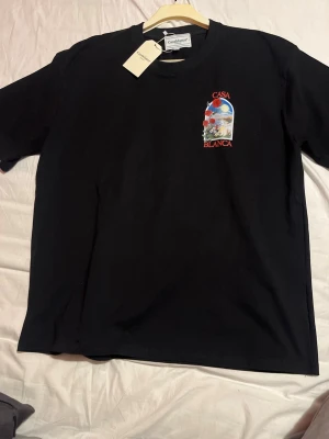 Svart t-shirt från Casablanca - Snygg helt ny svart t-shirt från Casablanca med prislapp och med liten färgglad grafik och texten 'Casa Blanca' på bröstet. Klassisk rund hals och korta ärmar. Perfekt för dig som gillar stilrena plagg med en twist. Tillverkad i mjuk bomull för skön känsla. Perfekt till sommaren. Och anledningen till att jag säljer dessa t shirts är för att dom är alldeles för stora för mig. 
