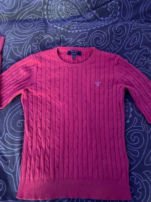 Rosa kabelstickad tröja från GANT - Säljer en snygg rosa kabelstickad tröja från GANT med rund halsringning och diskret logga broderad på bröstet. Tröjan har långa ärmar och ribbade muddar vid ärmslut och nederkant. Perfekt för dig som gillar klassisk och stilren design.