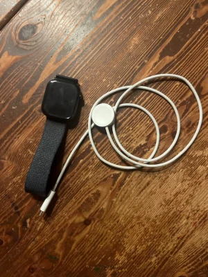 Svart Apple Watch med laddare - Snygg svart Apple Watch med rektangulär skärm och svart textilarmband. Klockan har en modern design och kommer med en vit magnetisk laddare. Perfekt för dig som vill ha koll på notiser, träning och hälsa direkt på handleden.