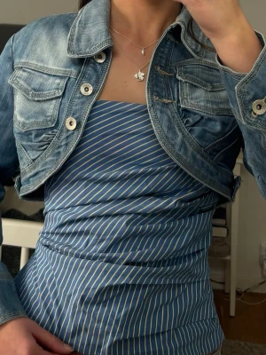 Kort jeansjacka i blå denim - Säljer en croppad jeansjacka i ljusblå denim med markerade sömmar och fyra fickor framtill. Jackan har en cool, figurnära passform och stora metallknappar. Perfekt att slänga över en klänning eller topp för en trendig look.