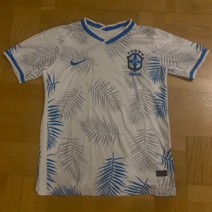 Brazil tröja - Säljer en vit Brasilien t-shirt från Nike med blå detaljer och palmbladsmönster över hela tröjan. T-shirten har korta ärmar, blå v-ringad krage och är tillverkad i ett lätt och skönt material som passar perfekt till fotboll eller träning. Pris ej hugget i sten